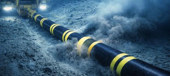 Subsea cable installation at the shoreline, with an excavator and workers guiding a black-and-yellow cable toward an offshore vessel, while an underwater trenching machine buries the cable on the seabed